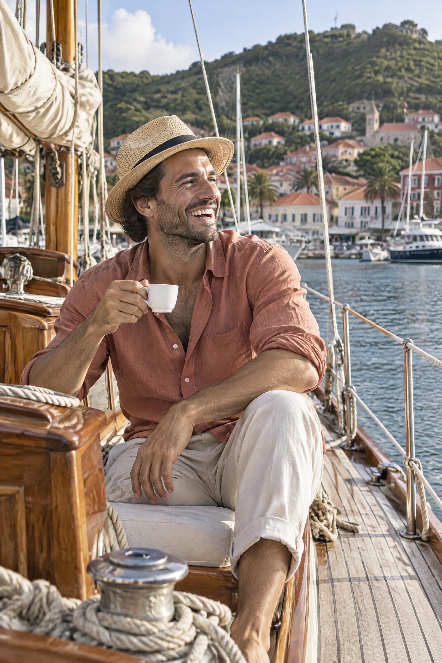 Un homme en chemise de lin terracotta et chapeau de paille rit sur le pont d’un voilier, le port derrière lui.