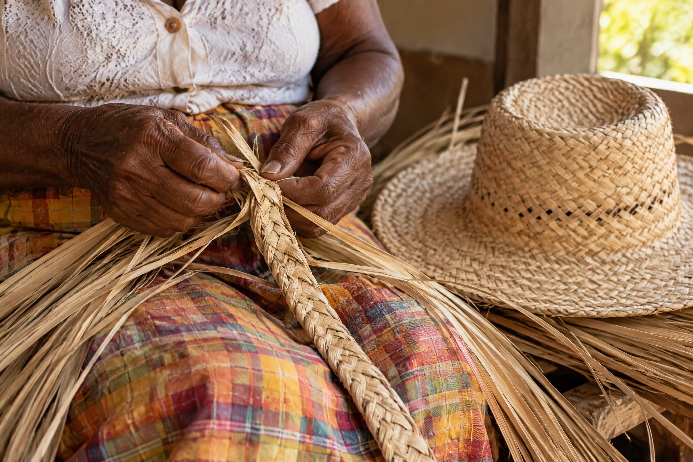 Des mains tressent une longue natte de paille ; un chapeau terminé est posé à côté.